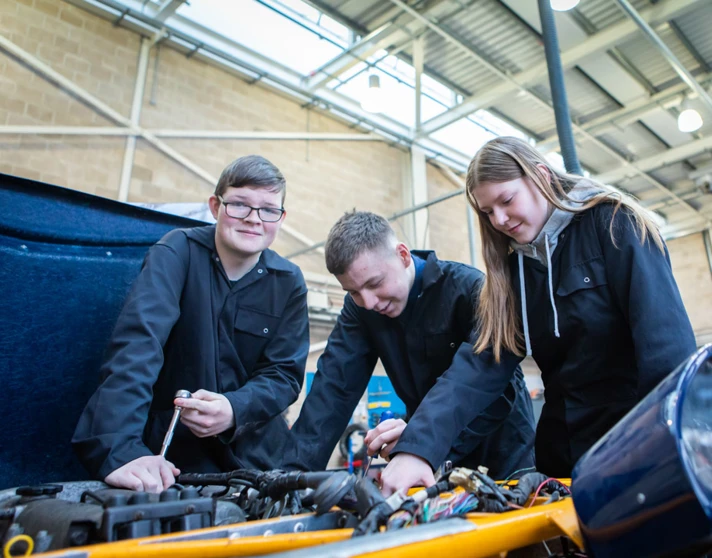 Three students working together under the hood of a car, using tools to inspect and repair an engine. Three students working together under the hood of a car, using tools to inspect and repair an engine.